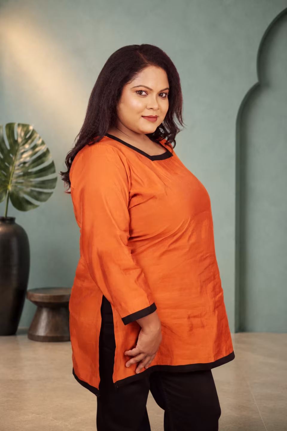 Side profile view of a woman wearing a solid orange square neck kurti showing the side slit and black contrast borders.