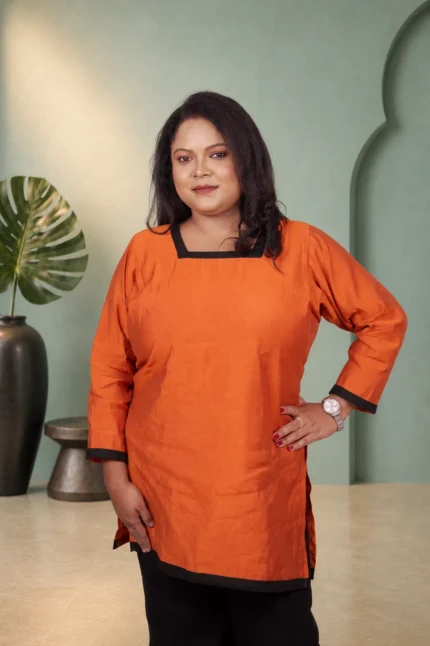 Woman posing with hand on hip in a solid orange square neck kurti with black contrast borders.