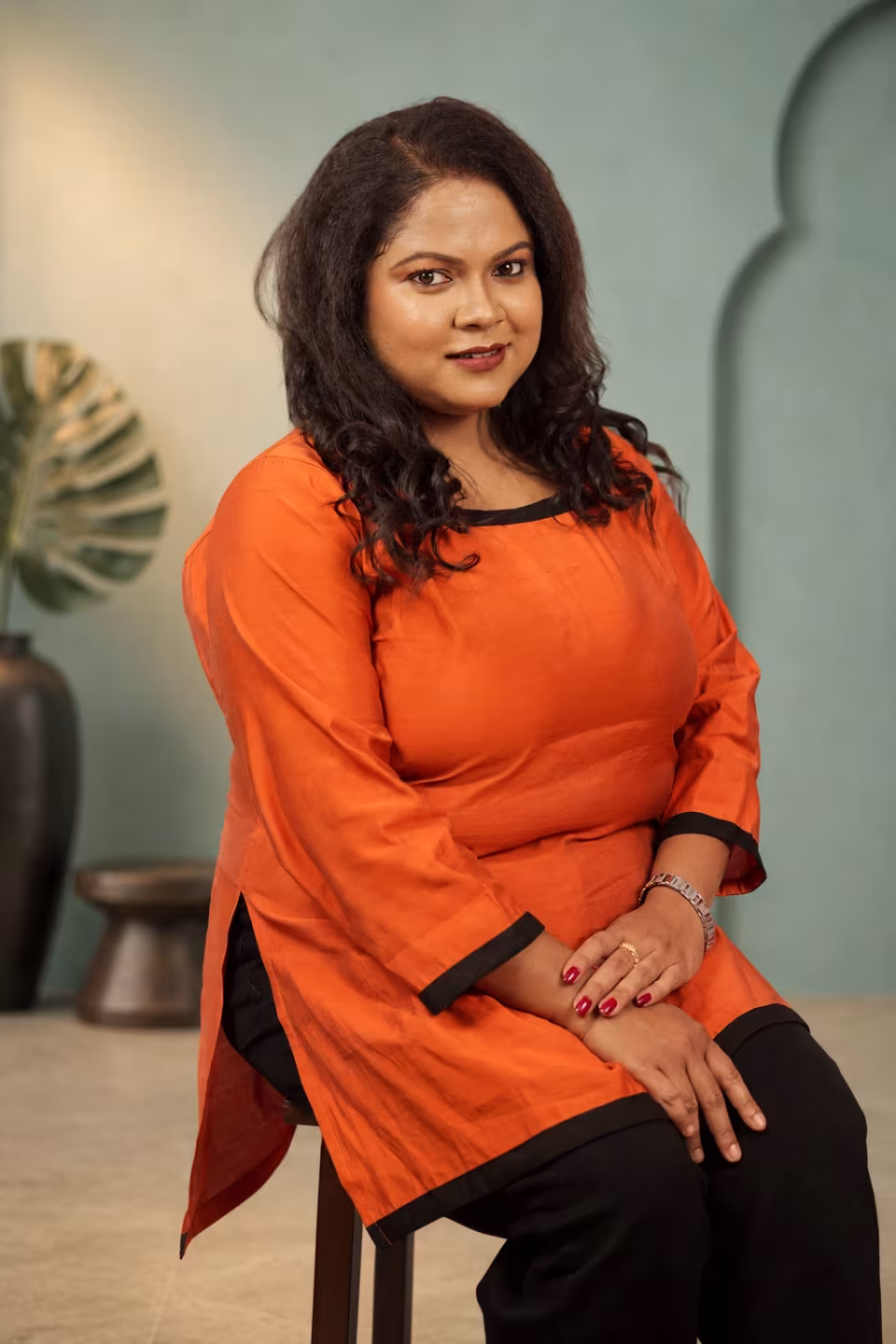 Woman sitting on a stool wearing a solid orange square neck kurti with black borders.